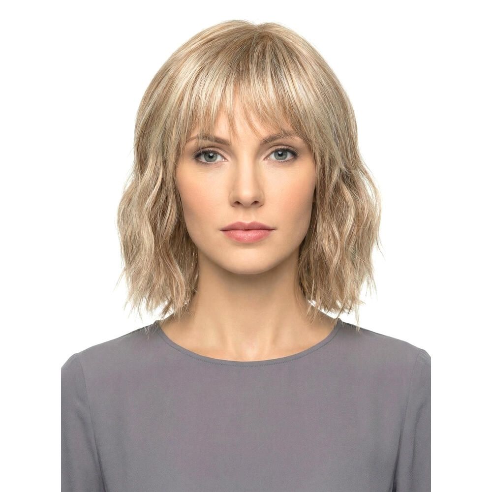 Blond gewellte Perücke mit Pony. Frontale Ansicht einer Frau mit grauer Bluse. Der Hintergrund ist weiß.