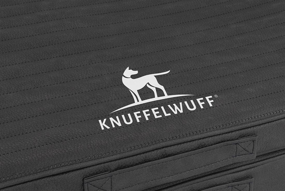 Detailansicht des Logos Knuffelwuff auf schwarzem, gestepptem Material. Tragegriff sichtbar.