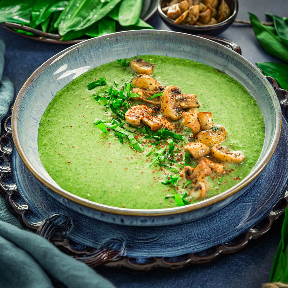 Grüne Suppe mit Pilzen und Kräutern in einer blauen Schüssel. Auf einem blauen Tablett. Dekoration mit Kräutern.