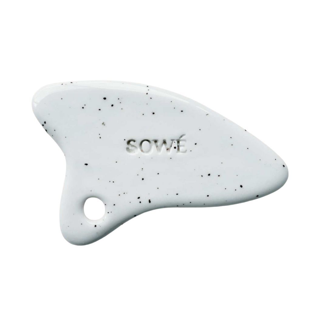 Weißes Gua Sha-Werkzeug mit schwarzen Sprenkeln. Keramik, mit Loch. Marke 'SOWÉ' eingeprägt.