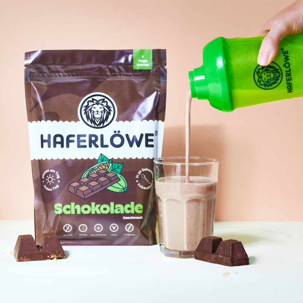 Schokoladen-Pulver von HAFERLÖWE wird in ein Glas gegossen. Schokoladenriegel liegen daneben. Grüner Shaker.