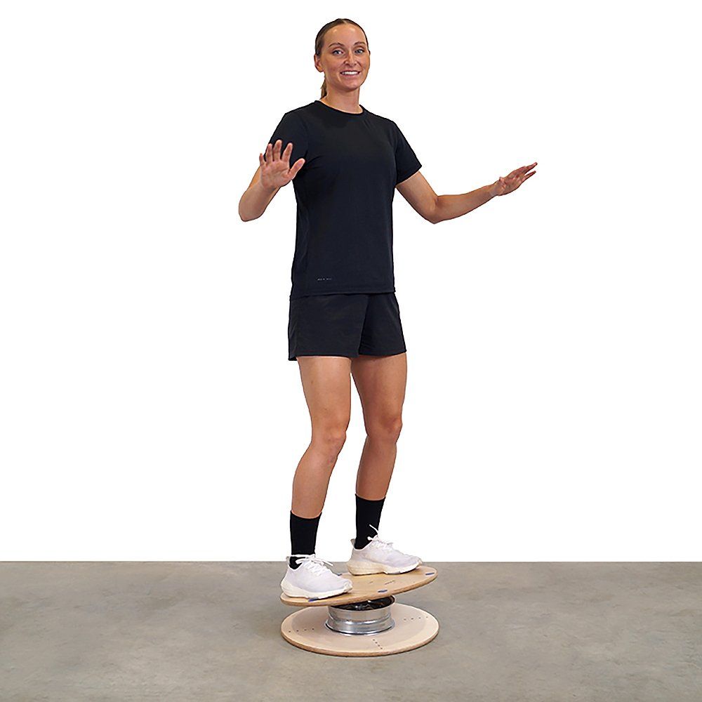 Person steht auf einem Pedalo Gleichgewichtstrainer. Das Gerät besteht aus Holz und Metall. Die Person balanciert mit ausgestreckten Armen.