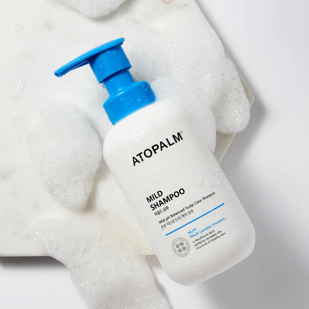 Weiße Flasche mit blauem Pumpkopf, umgeben von Schaum. Aufschrift ATOPALM, MILD SHAMPOO. Produkt für empfindliche Kopfhaut.