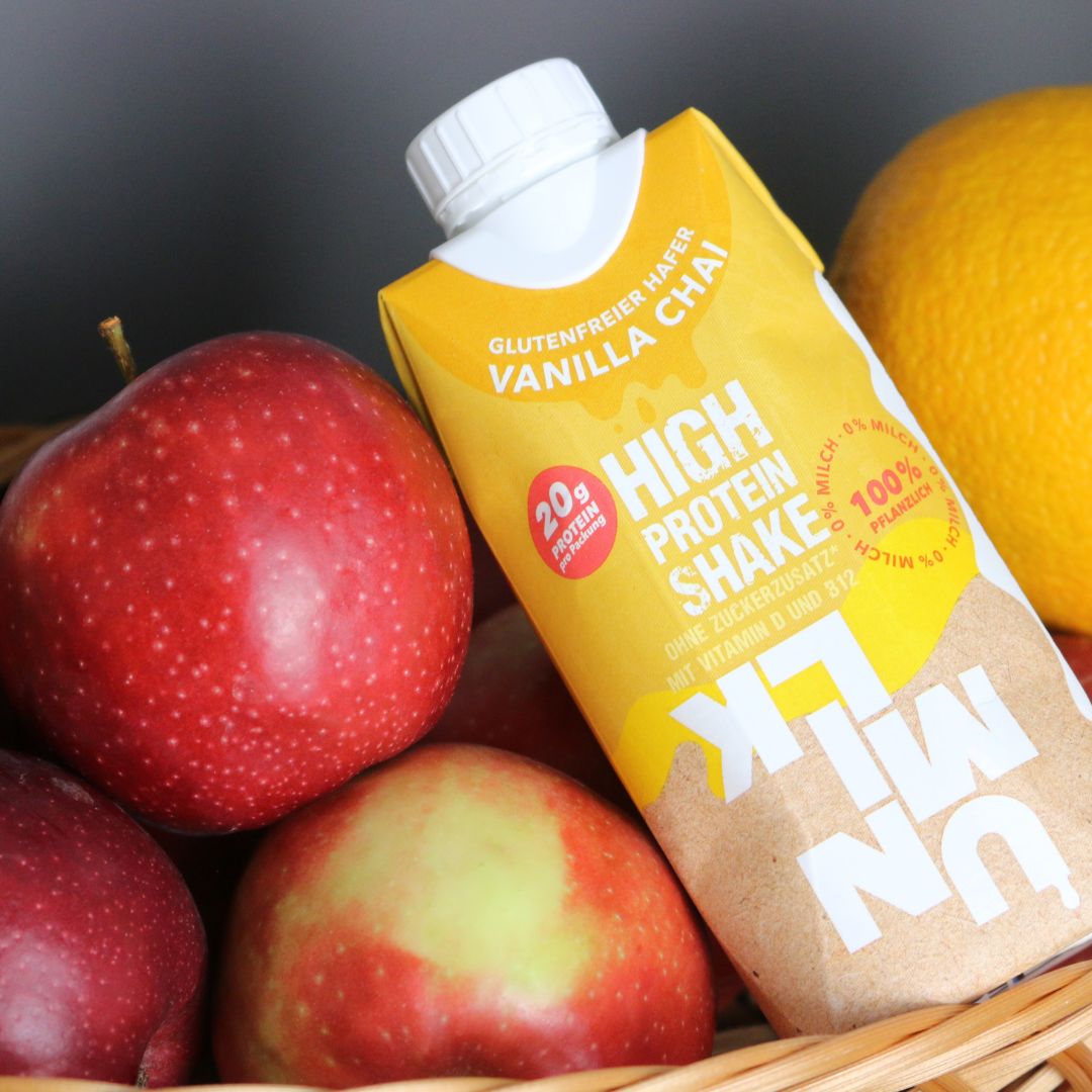 UNMILK High Protein Shake Vanilla Chai in einem Korb mit Äpfeln und einer Zitrone. Aufschrift: 20g Protein, glutenfrei, 100% pflanzlich.