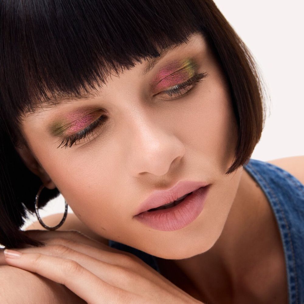 Nahaufnahme einer Frau mit geschminkten Augen. Lidschatten in Rosa und Grün. Kurze, dunkle Haare, blaue Jeansjacke.
