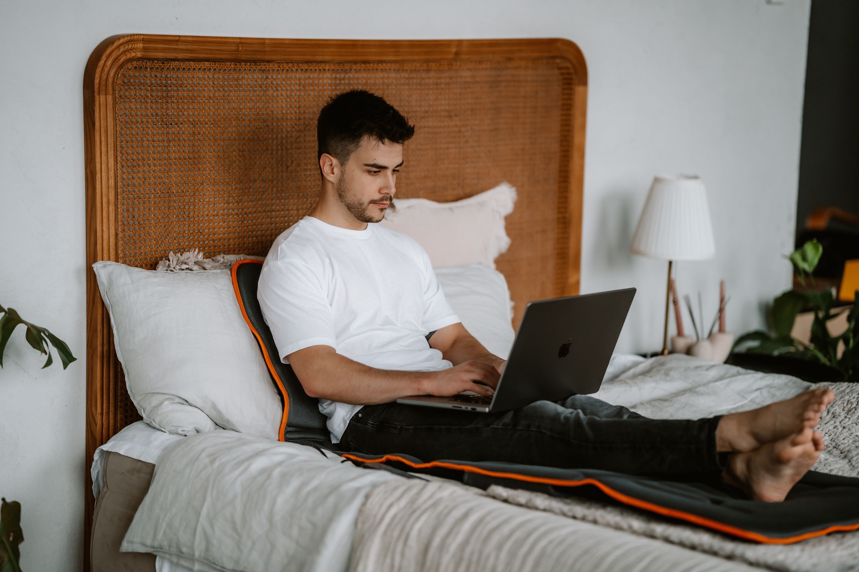 Mann sitzt auf einem Bett und arbeitet an einem Laptop. Unter ihm liegt eine graue Massagematte mit orangefarbenem Rand.