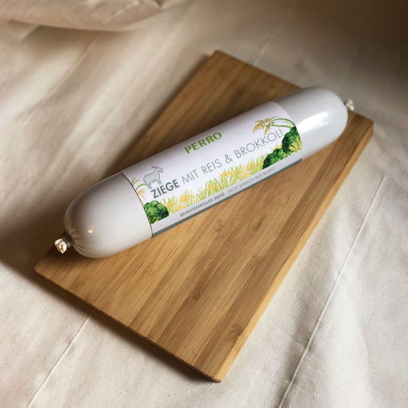 Hundewurst auf Holzbrett. Etikett: PERRO, Ziege mit Reis & Brokkoli. Produkt in weißer Hülle.
