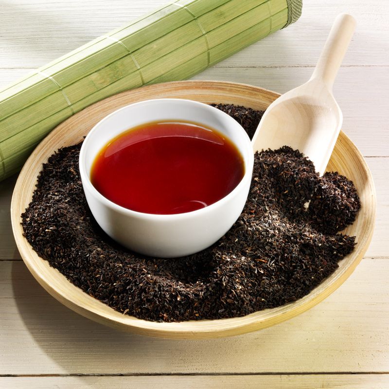 Tasse Tee in Holzschale mit losem Tee. Holzschaufel daneben. Bambusmatte im Hintergrund.