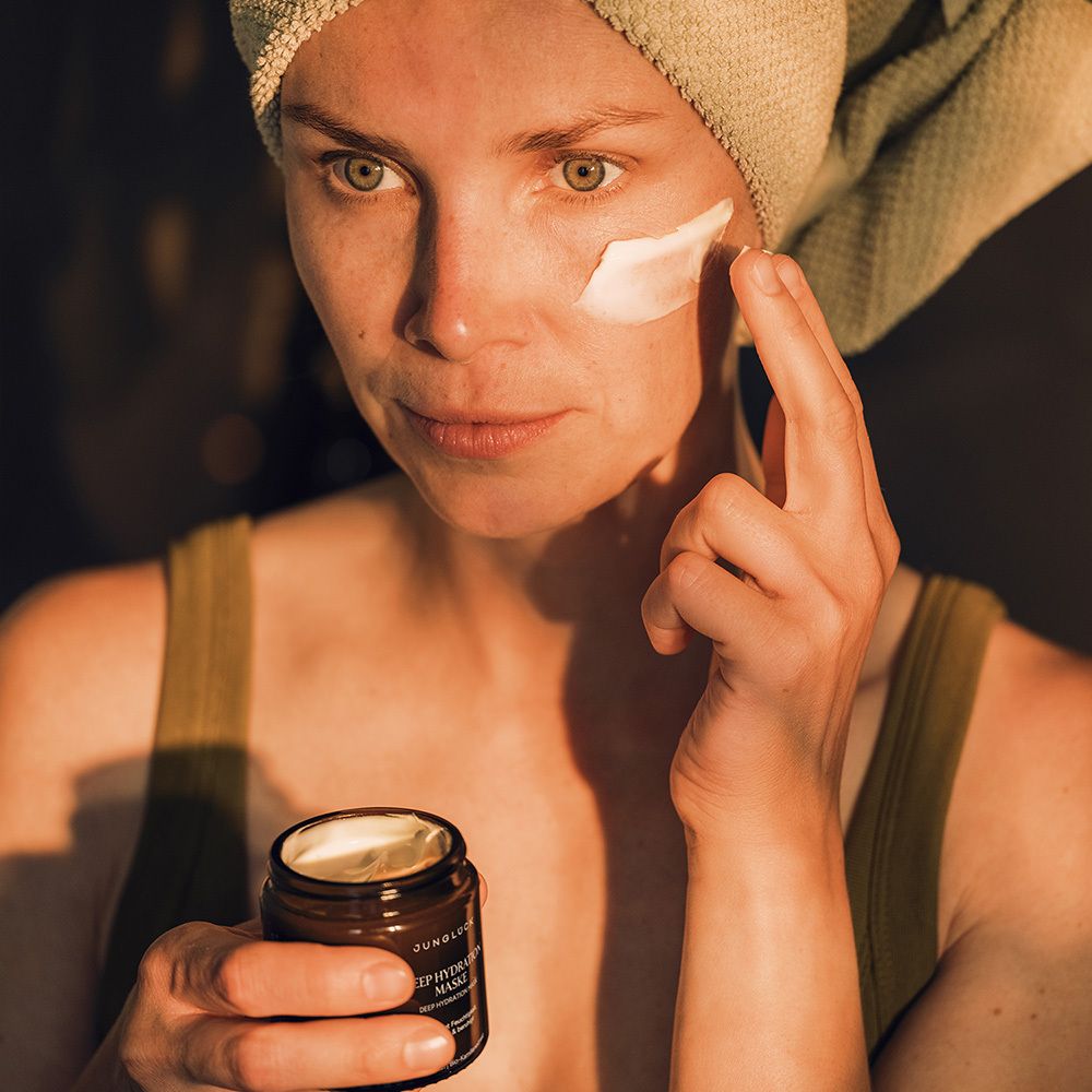 Frau mit Handtuch auf dem Kopf trägt Maske auf. Braunes Glas mit Produkt in der Hand. JUNGLÜCK Deep Hydration Maske.