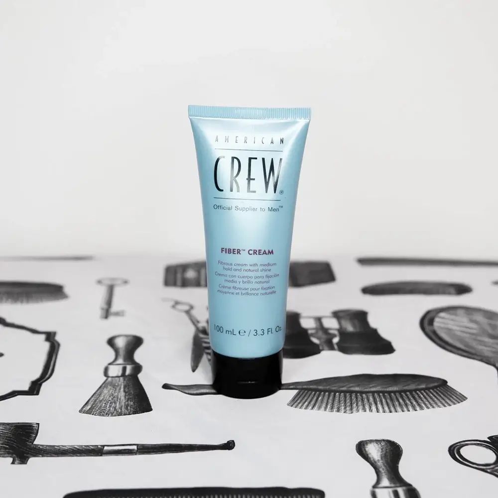 Türkisfarbene Tube mit schwarzem Deckel. Aufschrift: American Crew, Fiber Cream. Auf Tisch mit Friseursymbolen.