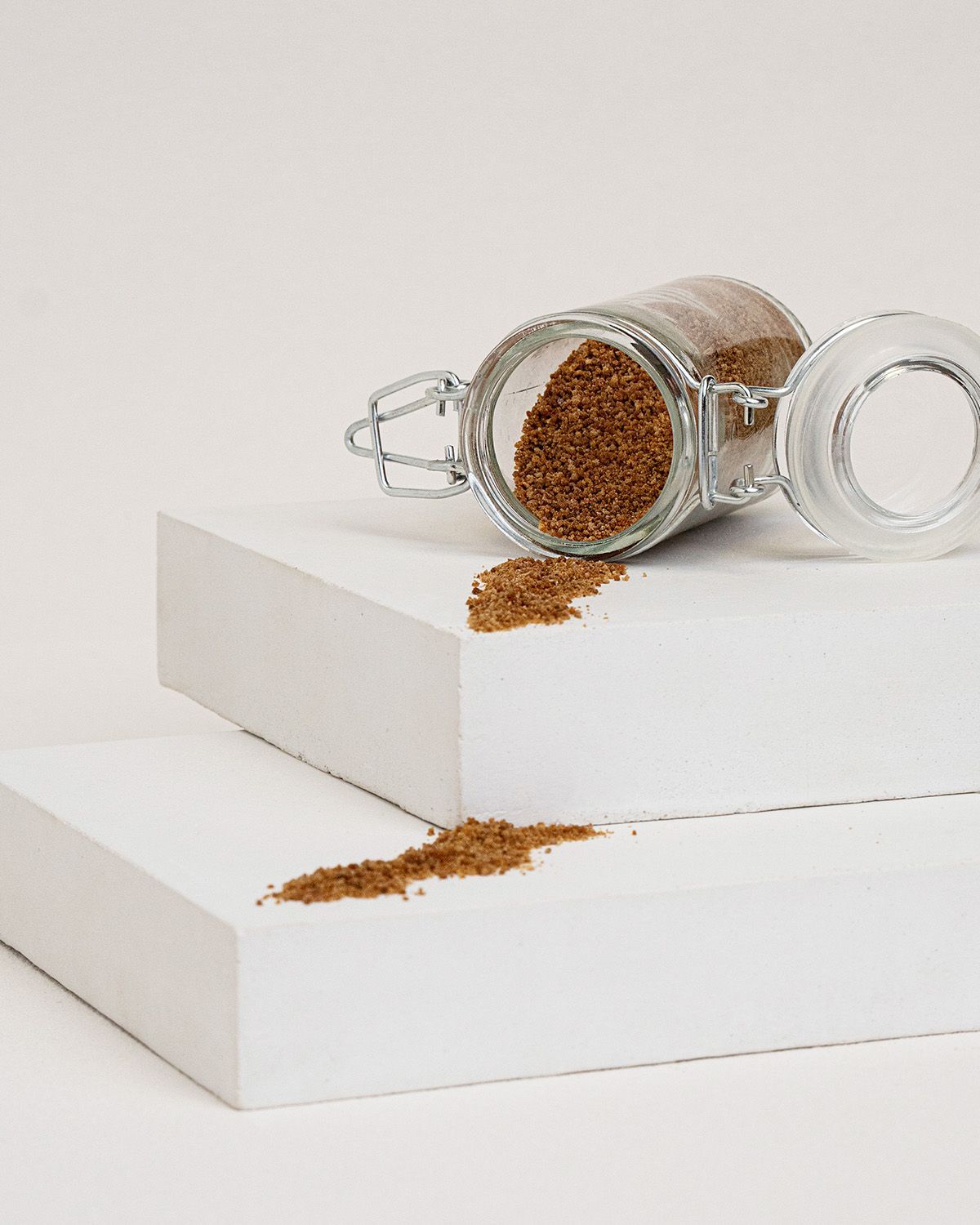 Glasbehälter mit braunem Zucker, daneben Zucker auf weißer Oberfläche. Deckel geöffnet.