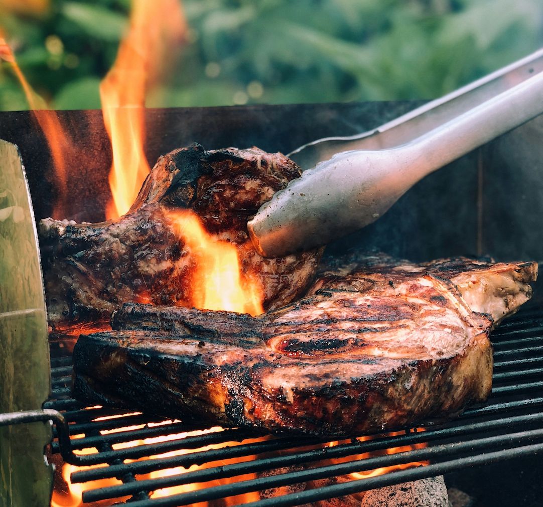 Grill mit zwei Steaks. Zange greift nach einem Steak. Flammen schlagen hoch.
