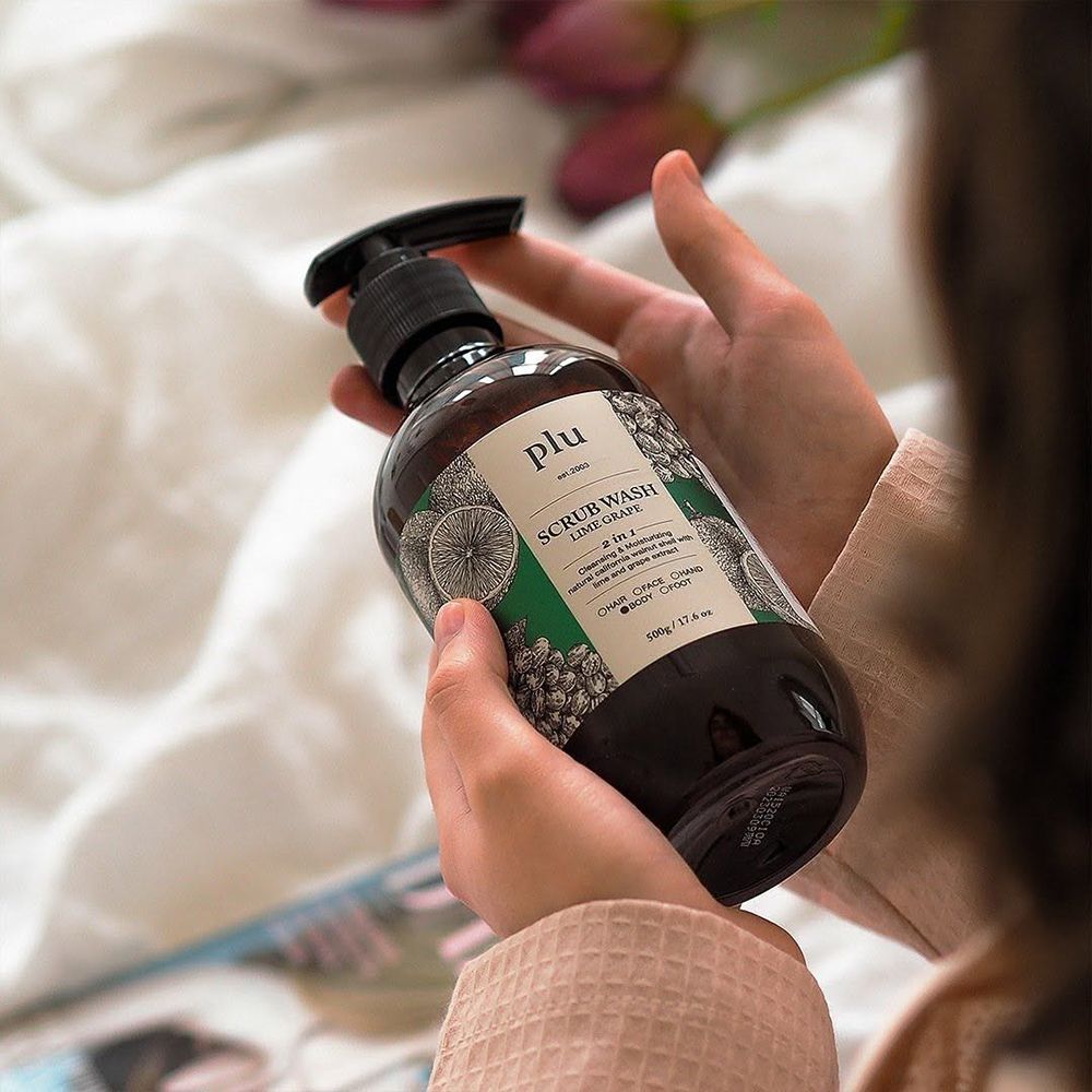 Person hält eine braune Flasche mit schwarzem Pumpverschluss. Etikett: plu, Scrub Wash Lime Grape. Helle Umgebung.
