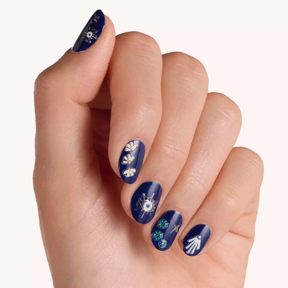 Hand mit lackierten Nägeln. Blaue Nägel mit goldenen und weißen Nagelstickern. Motive: Auge, Blüte, Muschel.