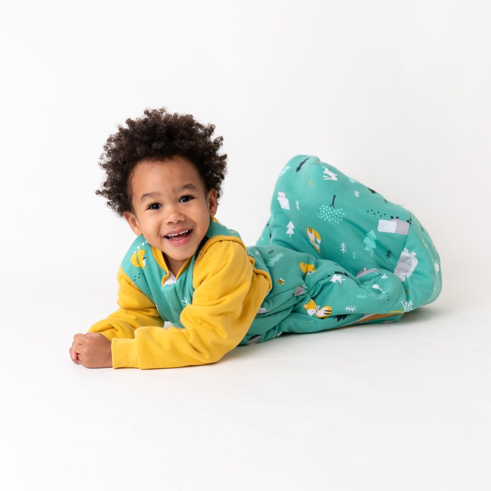 Kind liegt in grünem Schlafsack mit Waldtieren und gelben Ärmeln. Kind lächelt.