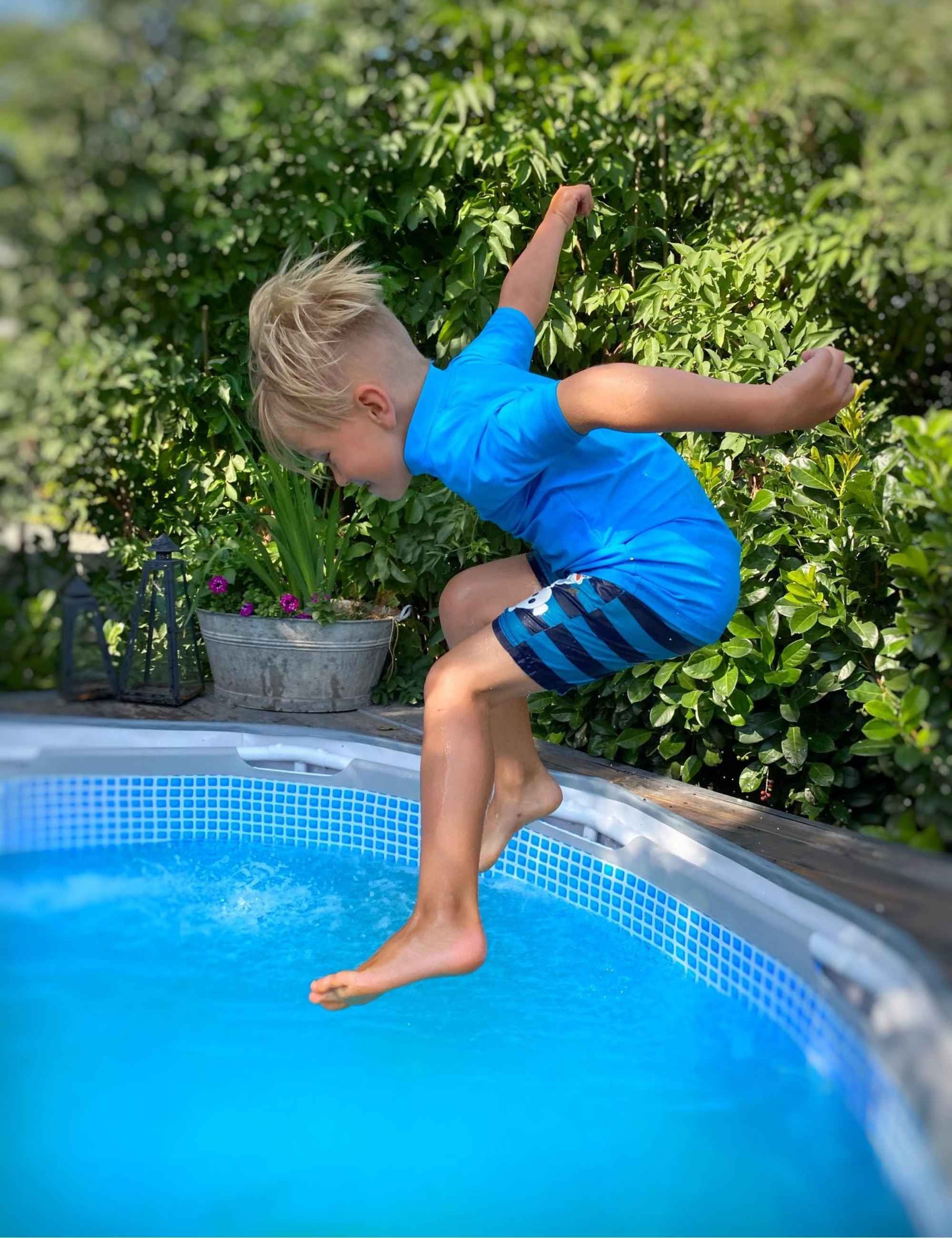 Kind springt in einen Pool. Trägt blaues Shirt mit IQ-UV-Logo und Badehose. Kurze Ärmel. Kind im Garten.