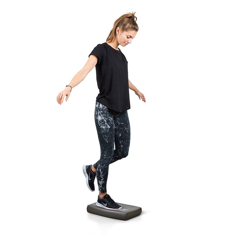Eine Frau balanciert auf einem Airex Balance-Pad Mini Lava. Sie trägt Sportkleidung und Turnschuhe.