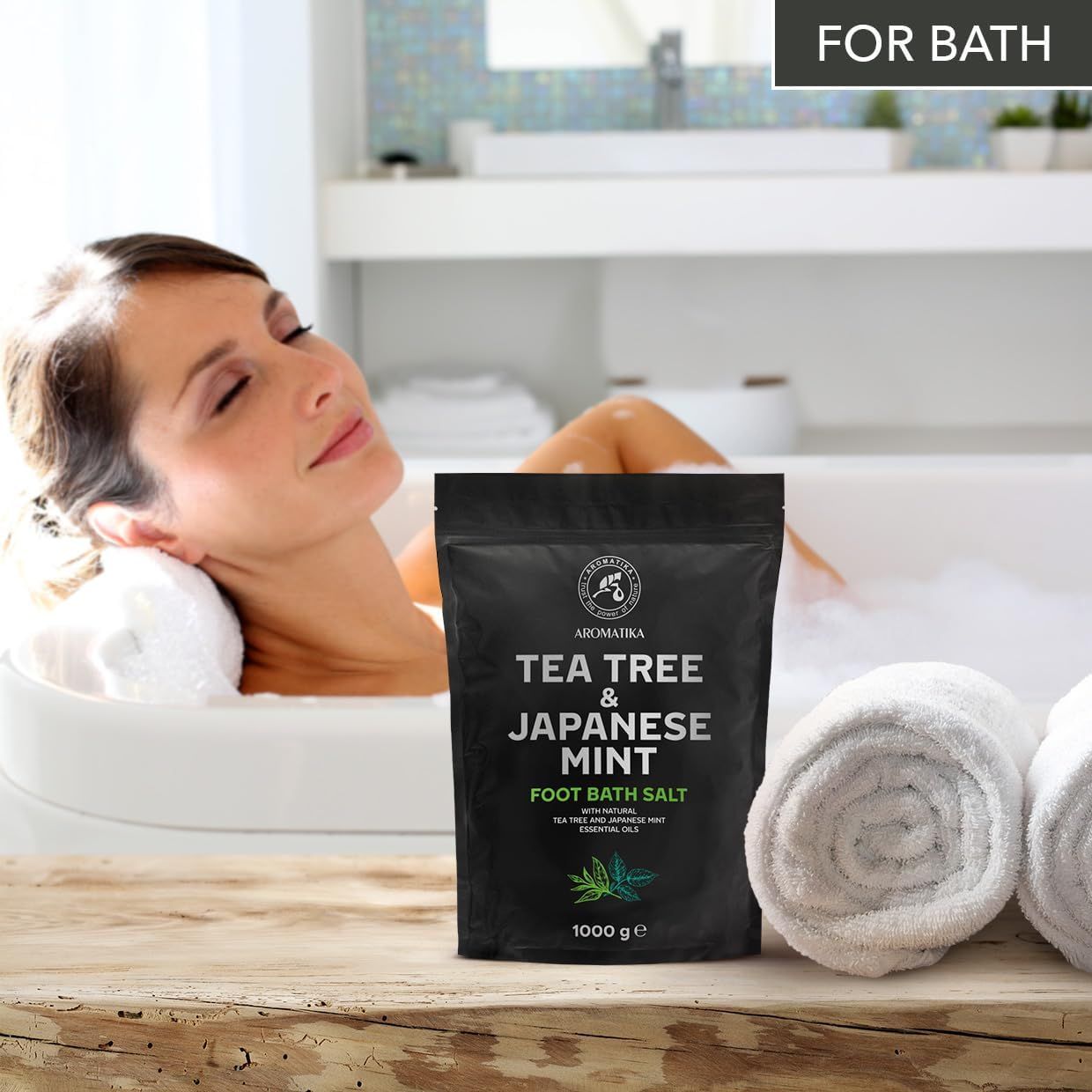 Frau in Badewanne mit Handtüchern. Schwarze Verpackung mit Produktnamen und Logo. Text: Tea Tree & Japanese Mint Foot Bath Salt. 1000 g.