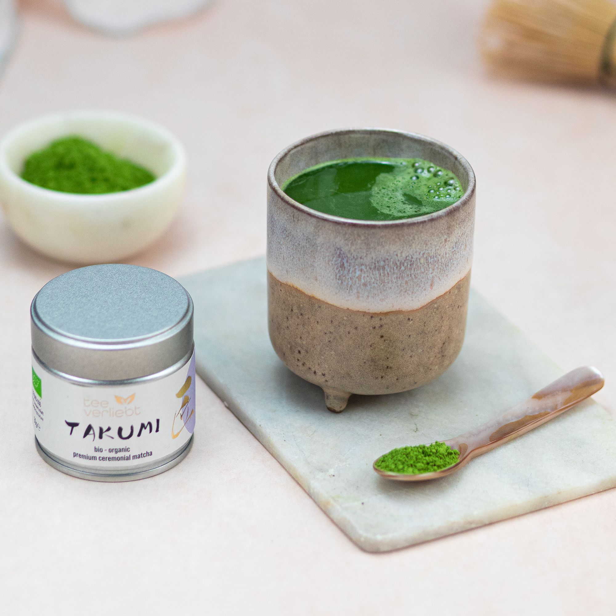 Dose und Tasse mit Matcha auf Marmorplatte. Löffel mit Matcha. Aufschrift: teeverliebt, TAKUMI, bio-organic, premium ceremonial matcha. Bio-Siegel.