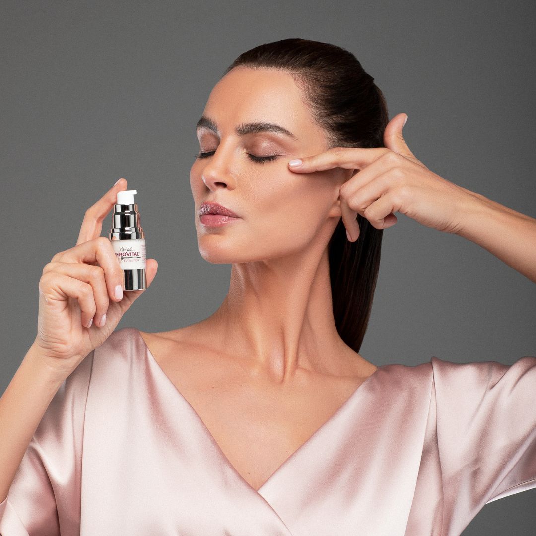 Frau hält Gerovital-Serum. H3 Evolution. Silberne Pumpe. Rosa Haut. Zeigt auf Augenpartie.