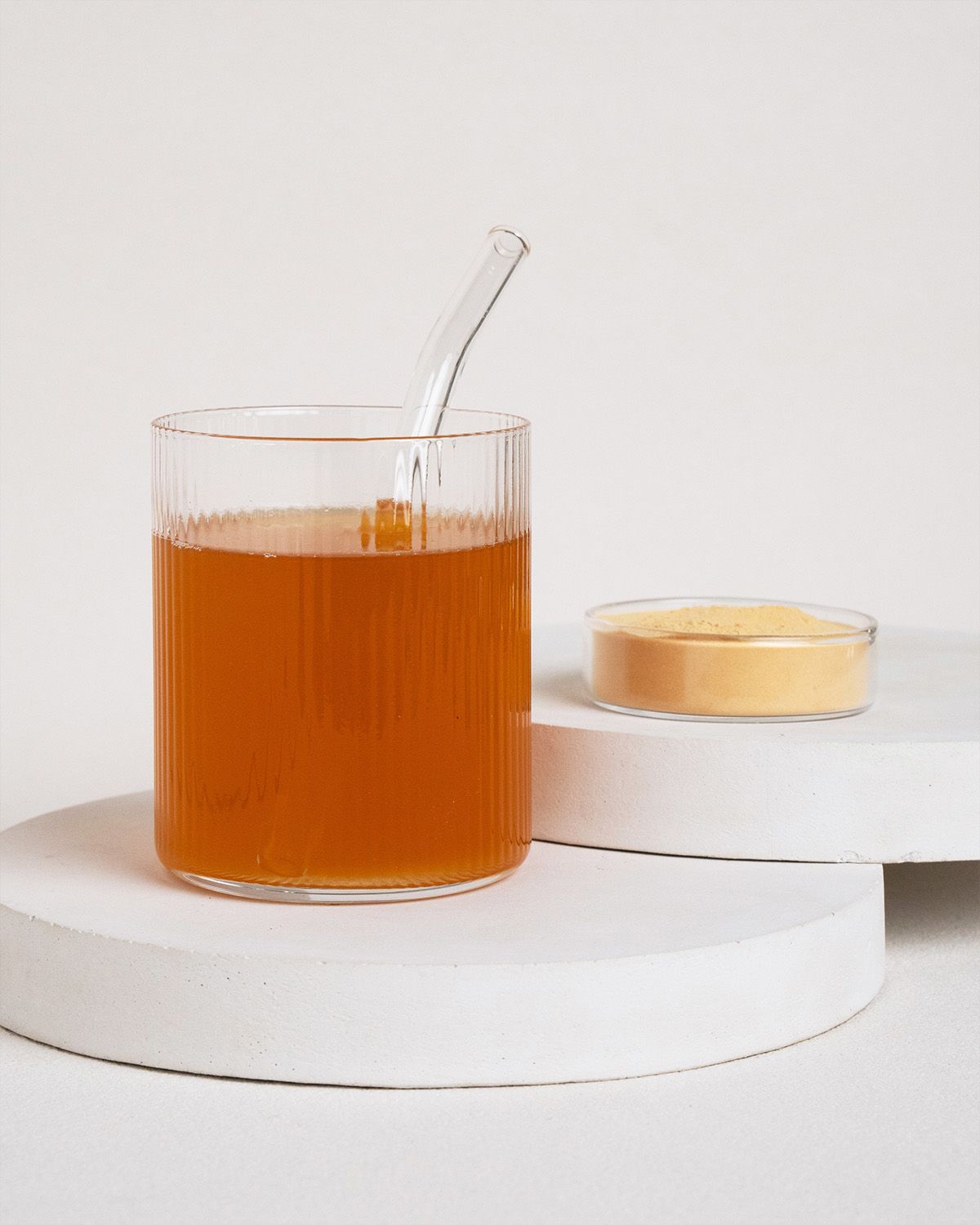 Glas mit Flüssigkeit und Trinkhalm, daneben Schale mit Pulver. Auf weißem Podest.