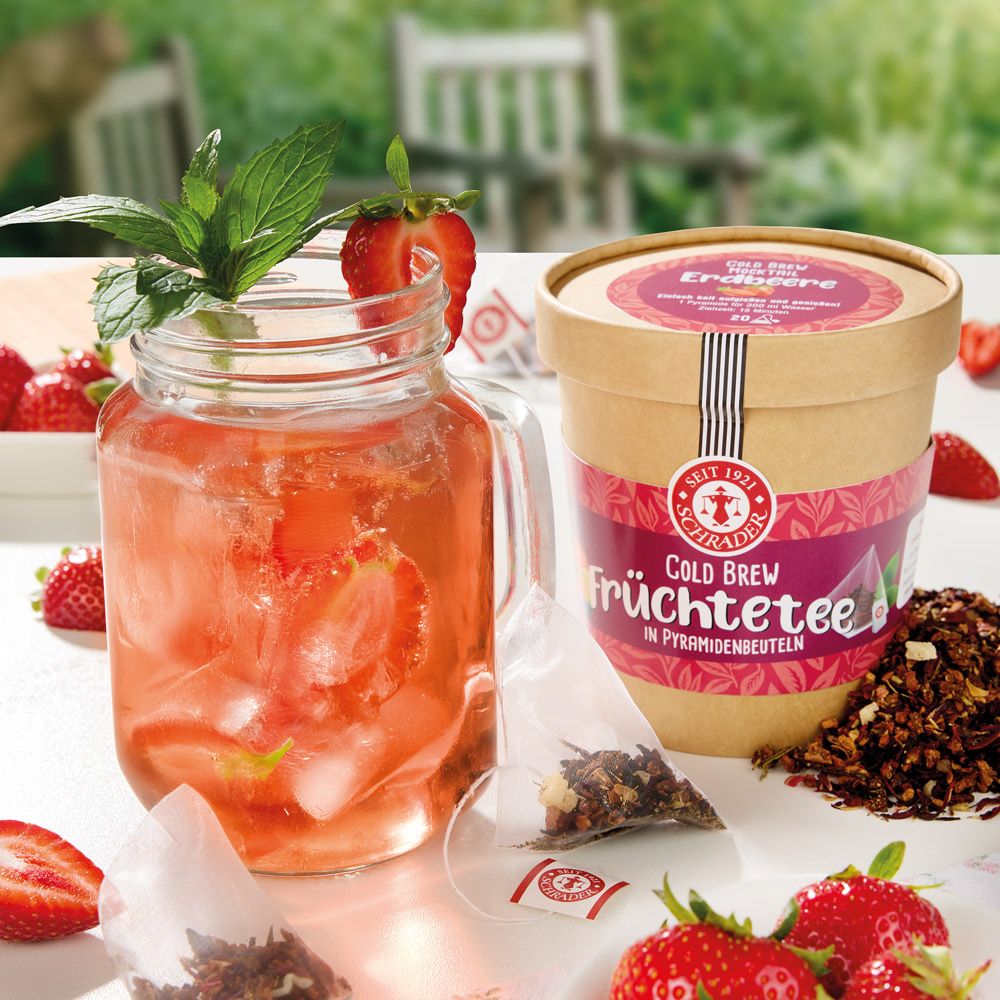 Glas mit Erdbeergetränk, daneben Dose Schrader Cold Brew Früchtetee Erdbeere. Erdbeeren und Teebeutel liegen daneben.