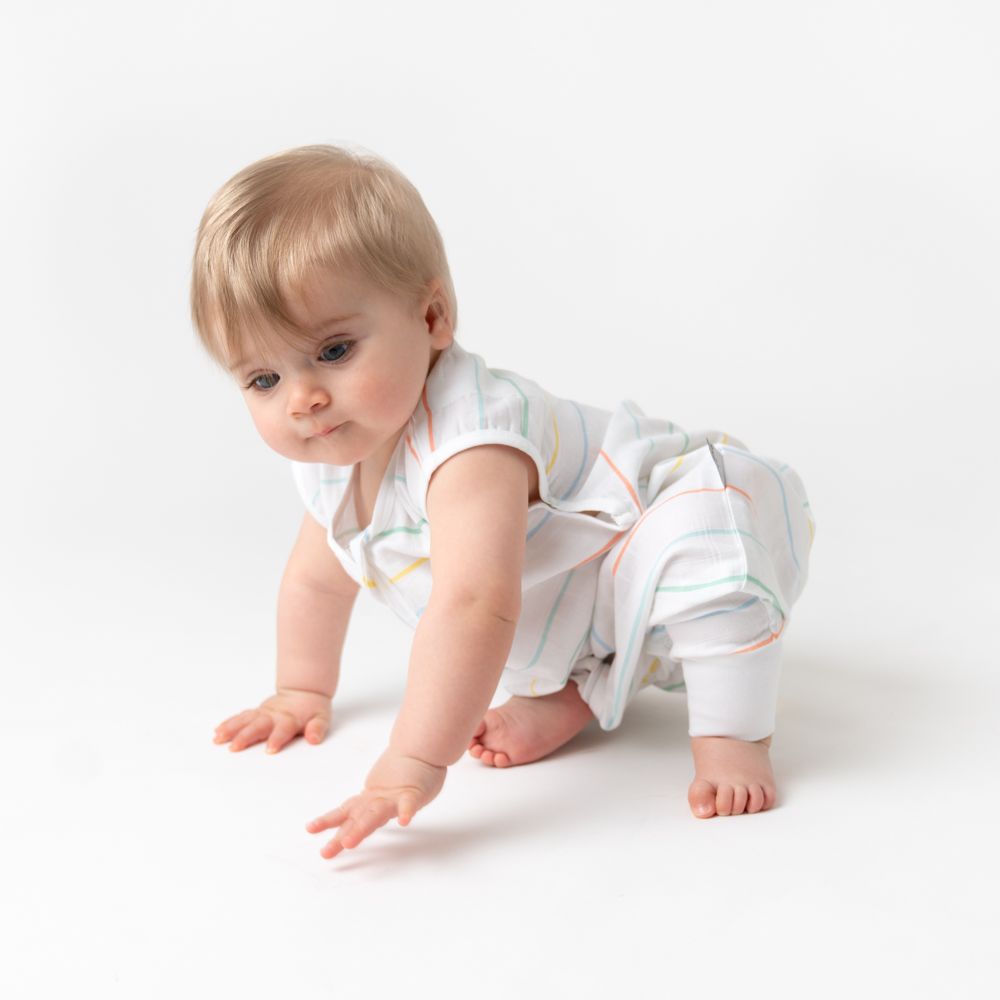 Baby krabbelt auf weißem Hintergrund. Trägt Schlafsack mit bunten Streifen und Füßen. Helle Haut, blonde Haare.
