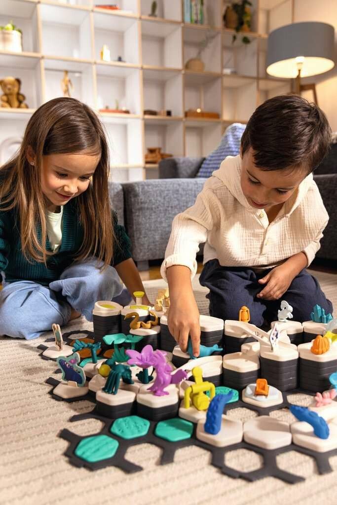 Due bambini giocano con il Ravensburger GraviTrax Junior Starter-Set XXL Planet. Il gioco è allestito sul pavimento, con vari elementi e personaggi. I bambini interagiscono con il gioco.