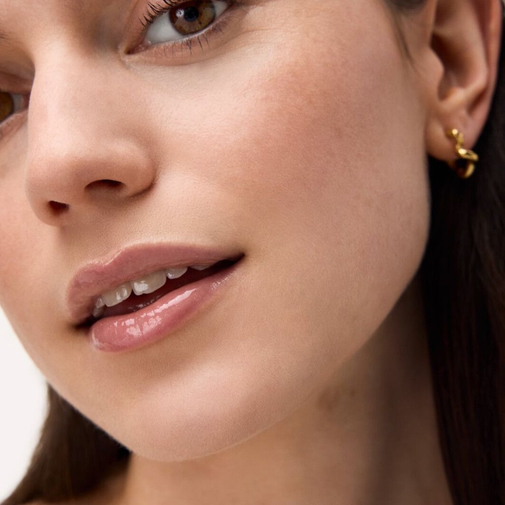 Nahaufnahme eines Gesichts mit glänzenden Lippen. Die Lippen haben einen rosafarbenen Farbton. Die Person trägt Ohrringe.