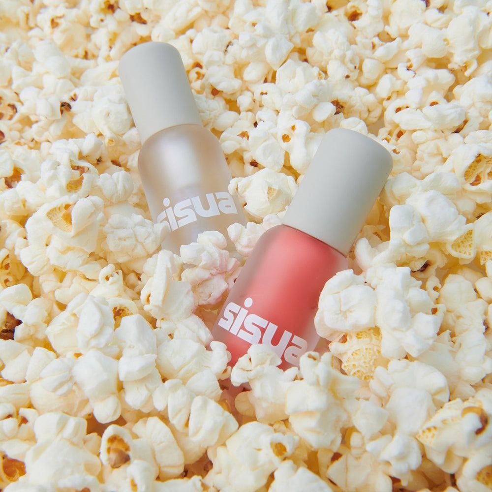 Zwei Flaschen mit Lipgloss, umgeben von Popcorn. Die Flaschen sind durchsichtig mit weißen Deckeln. Aufgedruckter Text: sisuia.
