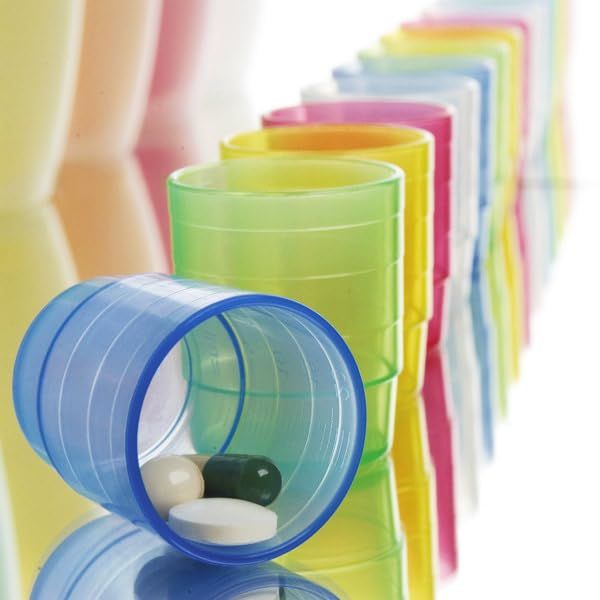 Reihe farbiger Becher, einer liegt mit Tabletten. Ohne Deckel. Auf glänzender Oberfläche.
