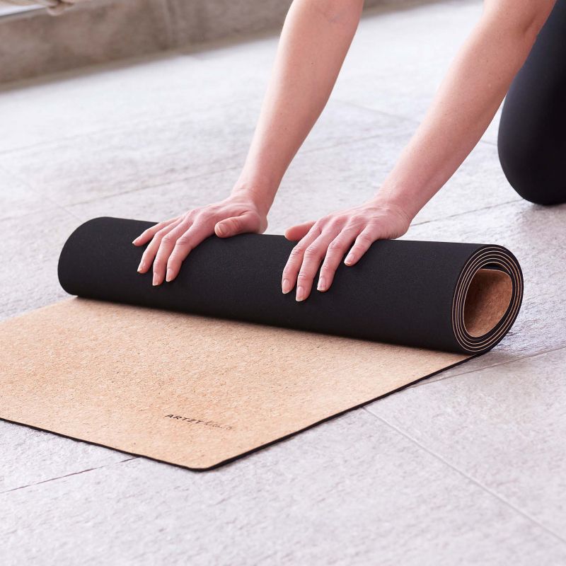 Person rollt Yogamatte aus Kork auf. Matte teilweise gerollt, hellbraun mit schwarzer Unterseite. ARTZT Vitality Logo.