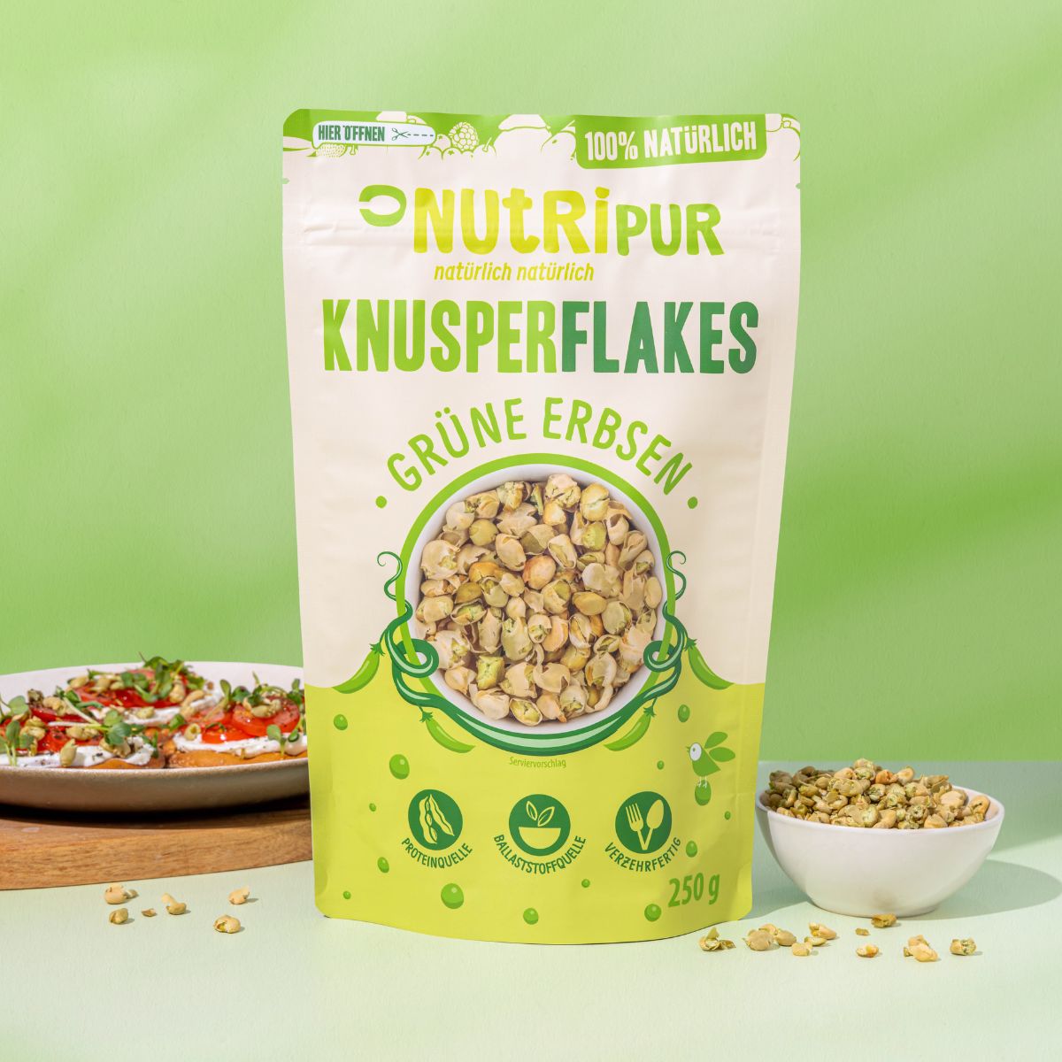 Verpackung von NutriPur Grüne Erbsen Knusperflakes. Aufschrift: Knusperflakes, Grüne Erbsen, 250g. Eine Schale mit Flakes daneben. Teller mit Essen.