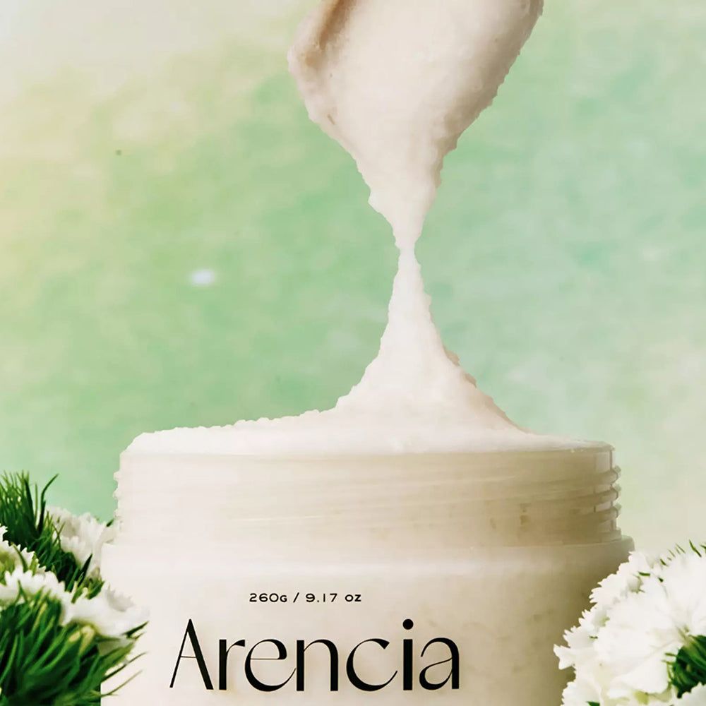 ARENCIA Peeling wird aus einem weißen Tiegel entnommen. Leichte Textur, umgeben von weißen Blüten.