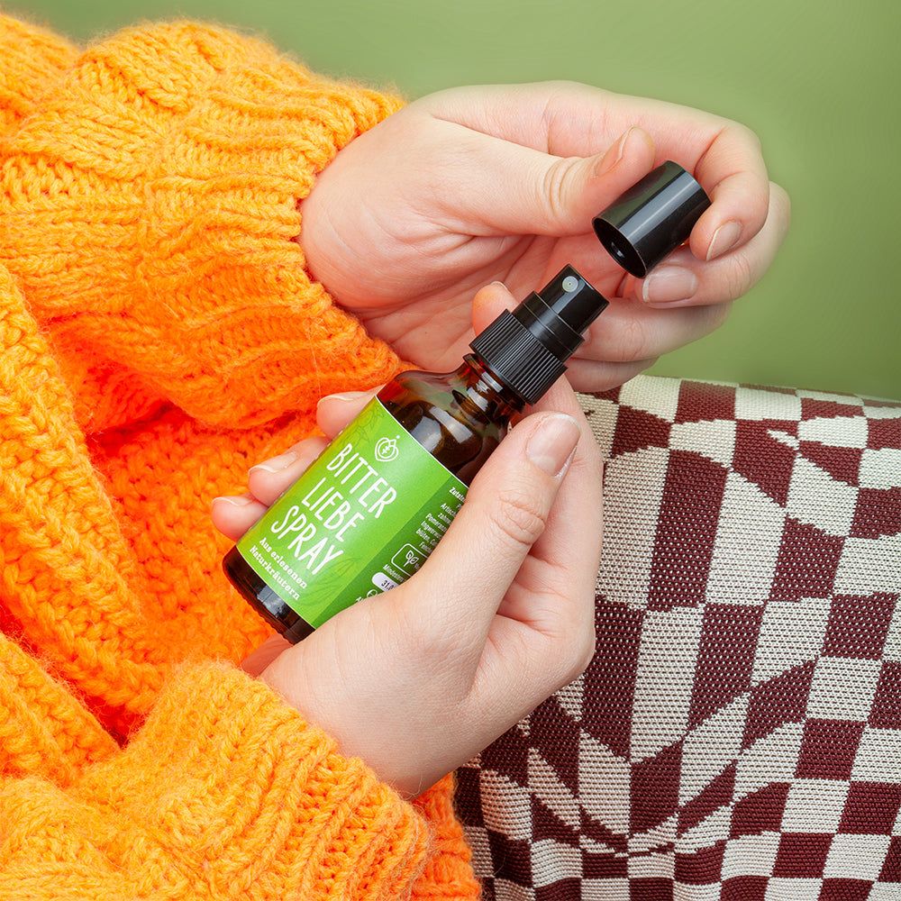 Hände halten eine braune Sprühflasche mit grünem Etikett. Aufschrift: BitterLiebe Spray. Schwarze Kappe wird geöffnet.