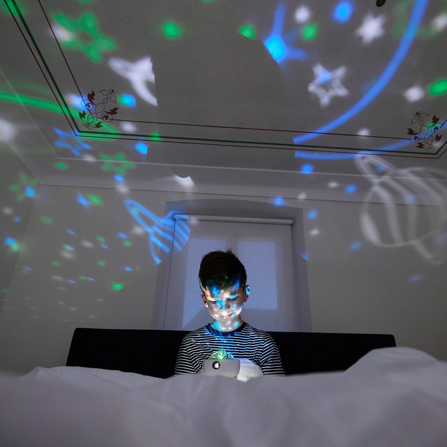 Kind im Bett, beleuchtet von Sternenprojektion. Projektor in der Hand. Sterne und Planeten an der Decke.