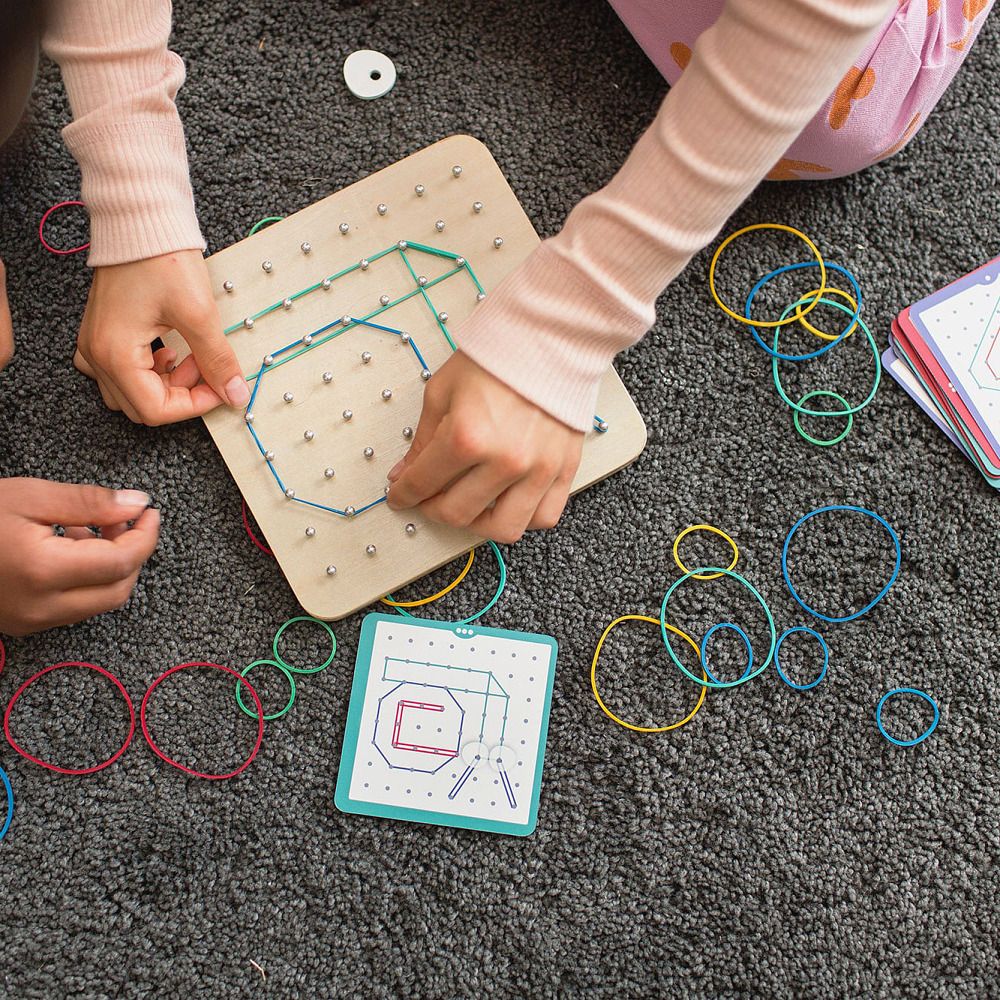Kind spielt mit Geoboard. Holzbrett mit Gummibändern. Karten und Gummibänder liegen daneben.