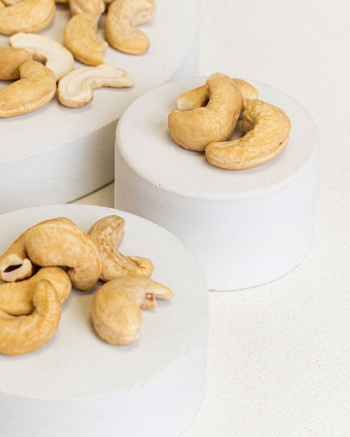 Cashewkerne auf weißen Podesten. Helle, beige Nüsse in Halbmondform. Einige Kerne sind angeschnitten.
