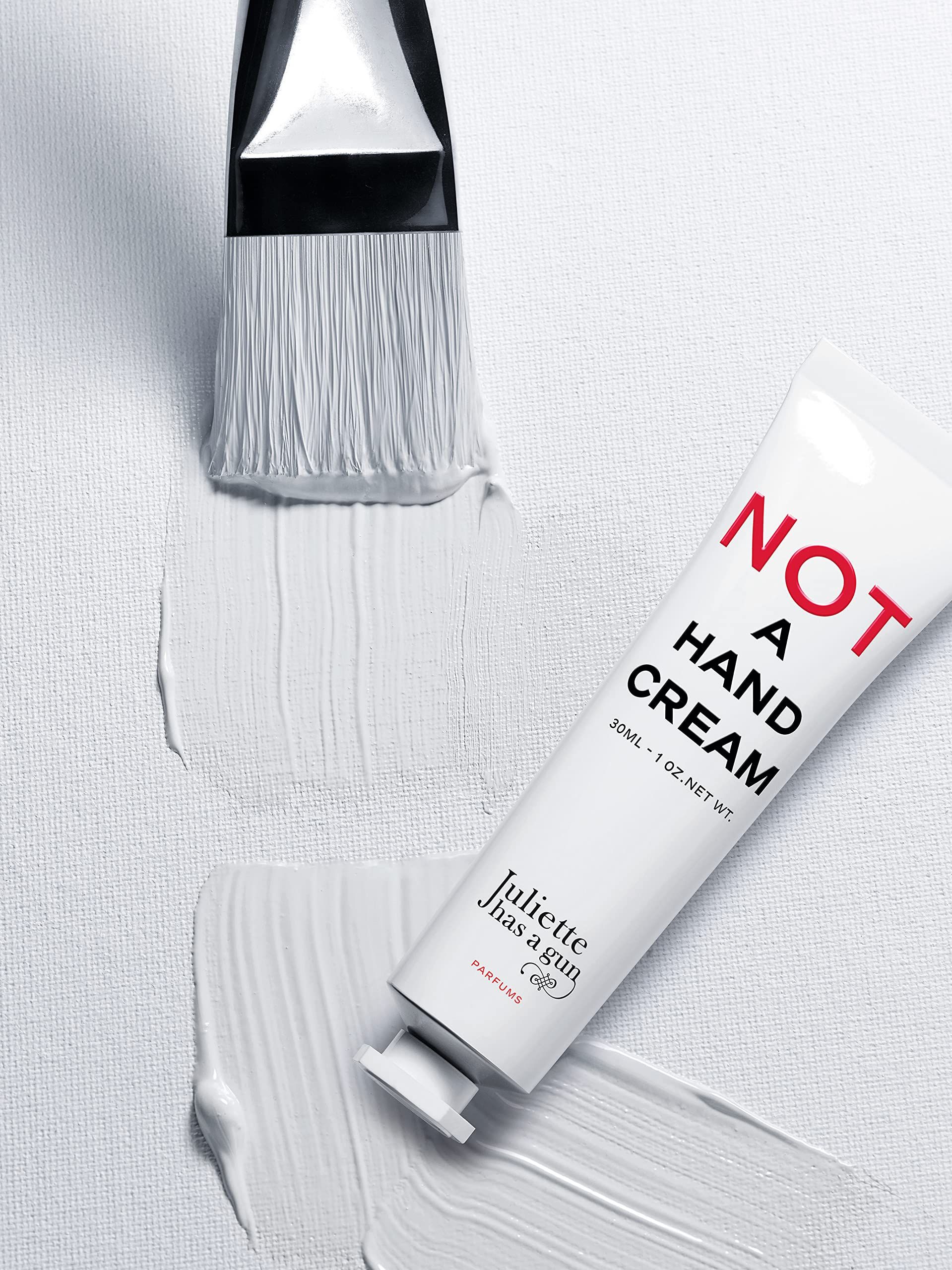 Weiße Handcreme-Tube mit rotem Schriftzug 'NOT A HAND CREAM'. Pinsel mit weißer Farbe daneben. Marke: Juliette Has a Gun.