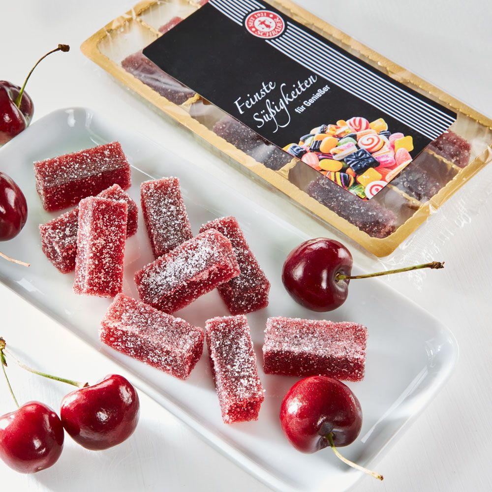 Rote Gelee-Stücke auf weißem Teller, daneben Kirschen. Gelee-Stücke mit Zucker bestreut. Verpackung im Hintergrund.