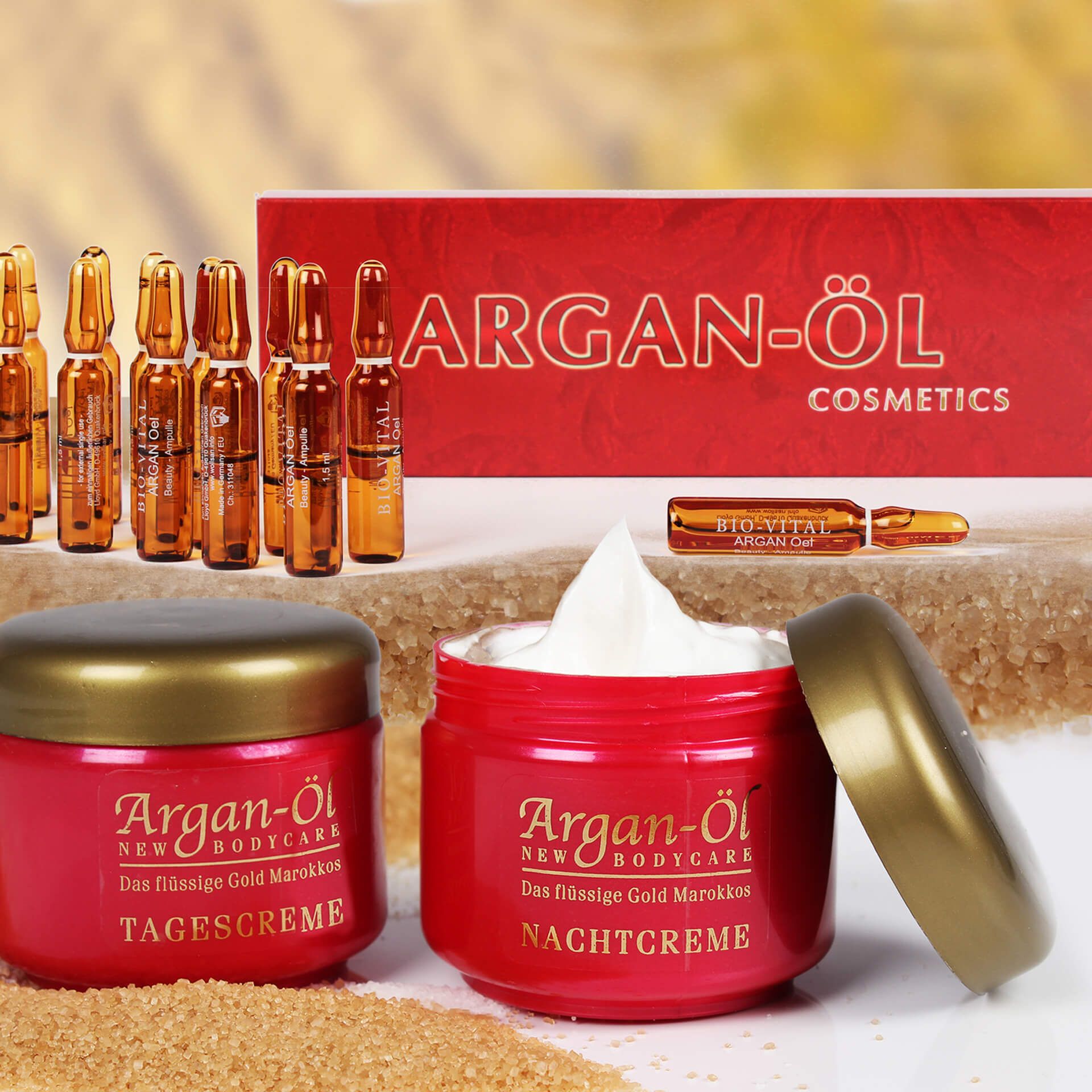 Rotes Tiegel mit goldfarbenem Deckel, daneben offener Tiegel mit Creme. Ampullen und Karton mit Schriftzug Argan-Öl.