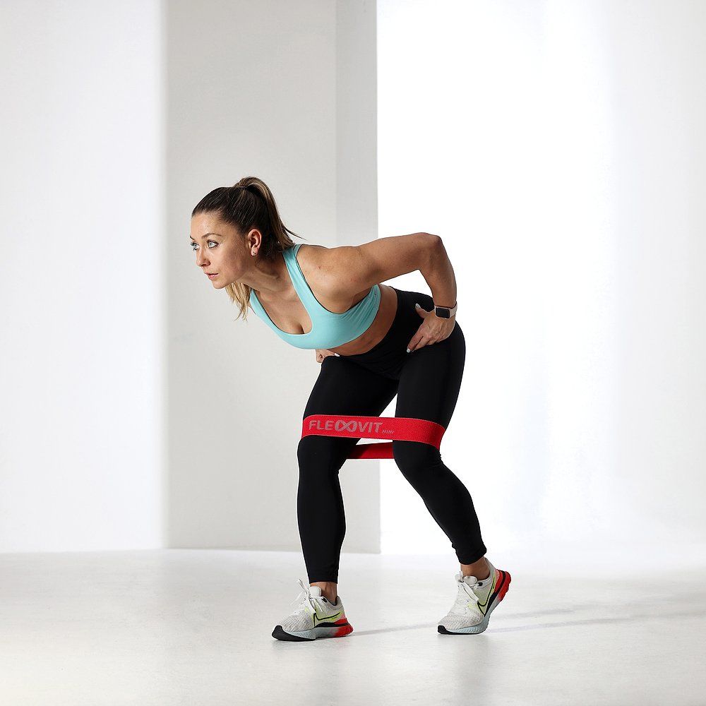 Frau mit rotem FLEXVIT Mini-Band um die Beine. Sie beugt sich nach vorne. Schwarze Leggings und Sport-BH.