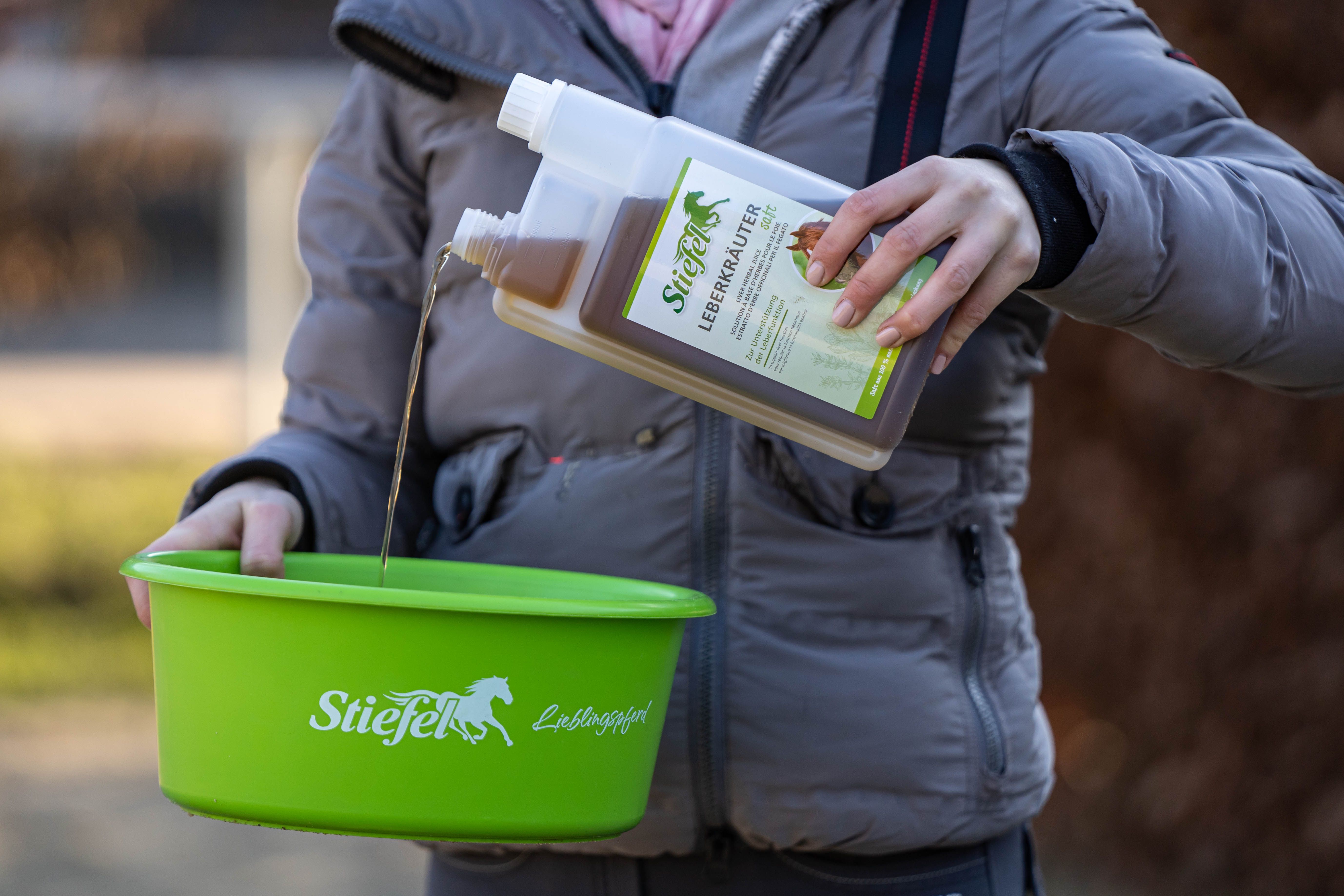 Person gießt Stiefel Leberkräutersaft aus einer Flasche in eine grüne Schüssel. Produktname und Logo sind sichtbar.