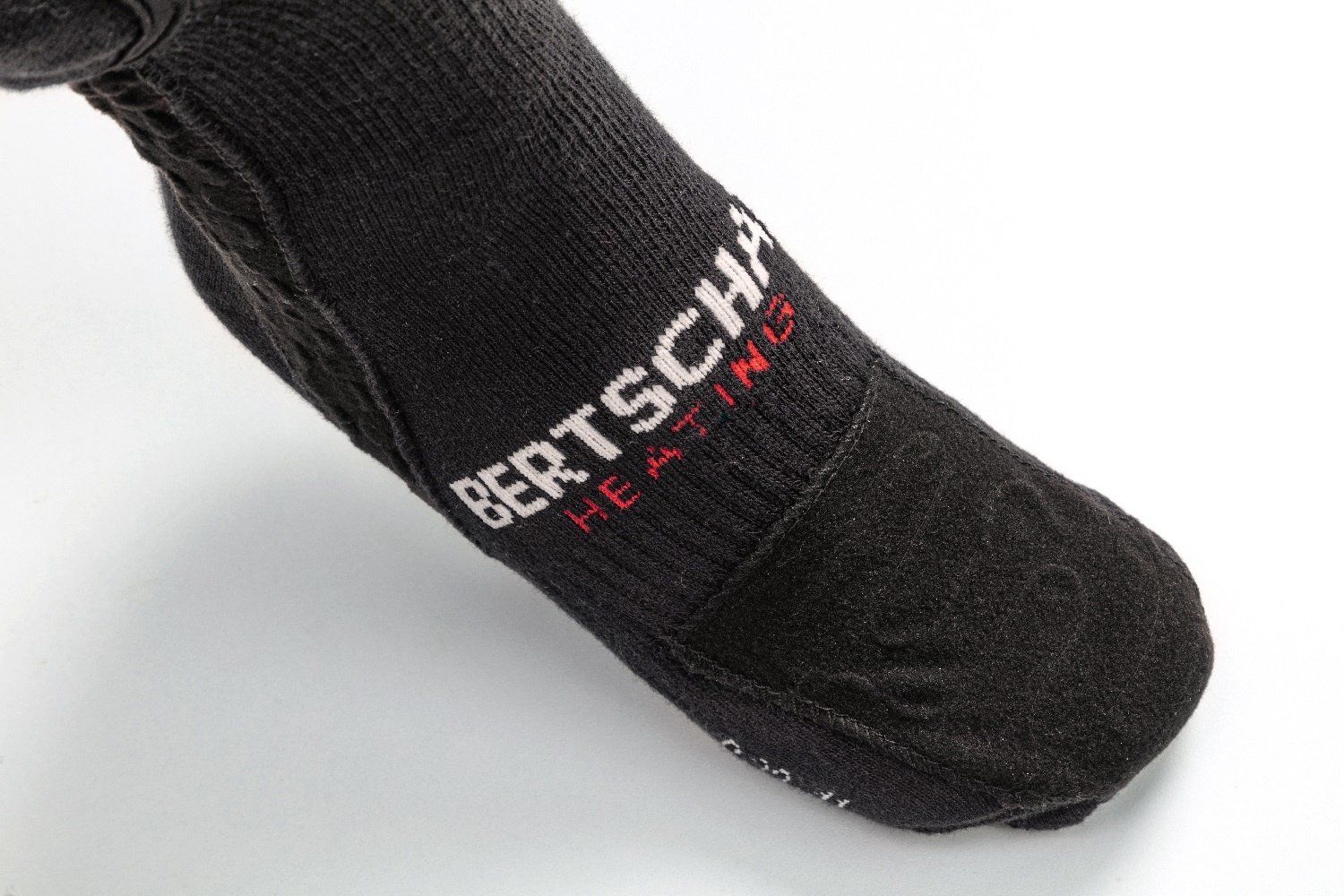 Nahaufnahme der Sohle einer schwarzen BERTSCHAT® Socke. Das Logo und ein rutschfestes Muster sind sichtbar.