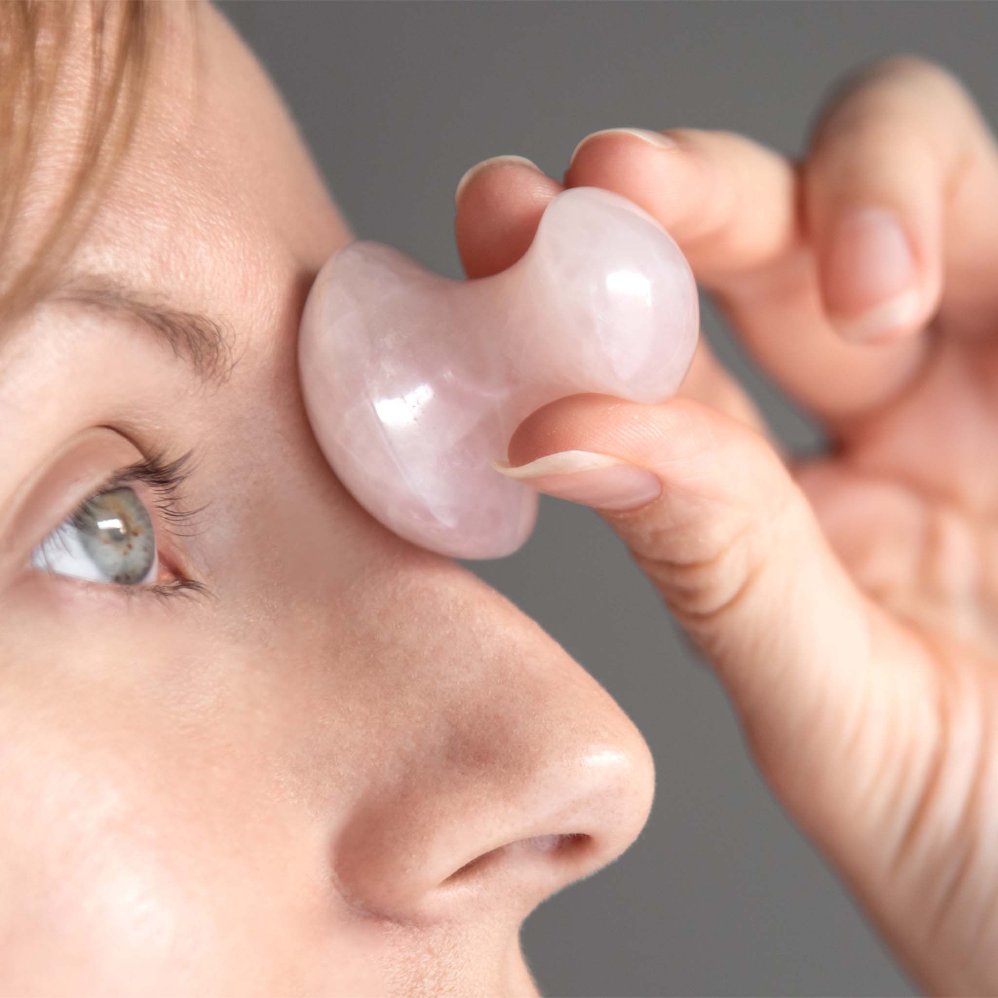Persoon houdt rozenkwarts Gua Sha in paddenstoelvorm bij het oog. Grijze achtergrond.