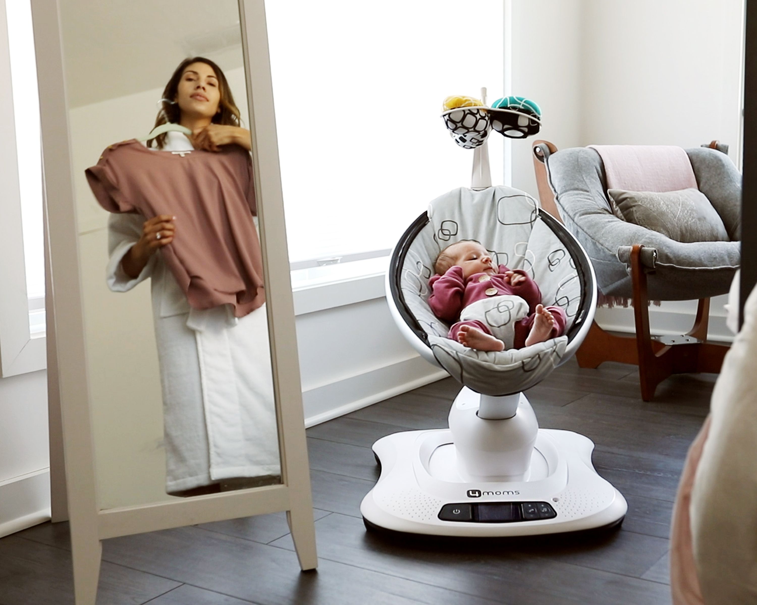Baby in Babywippe. Grauer Sitz mit Muster, Spielzeug. Frau mit Spiegel. Innenraum.