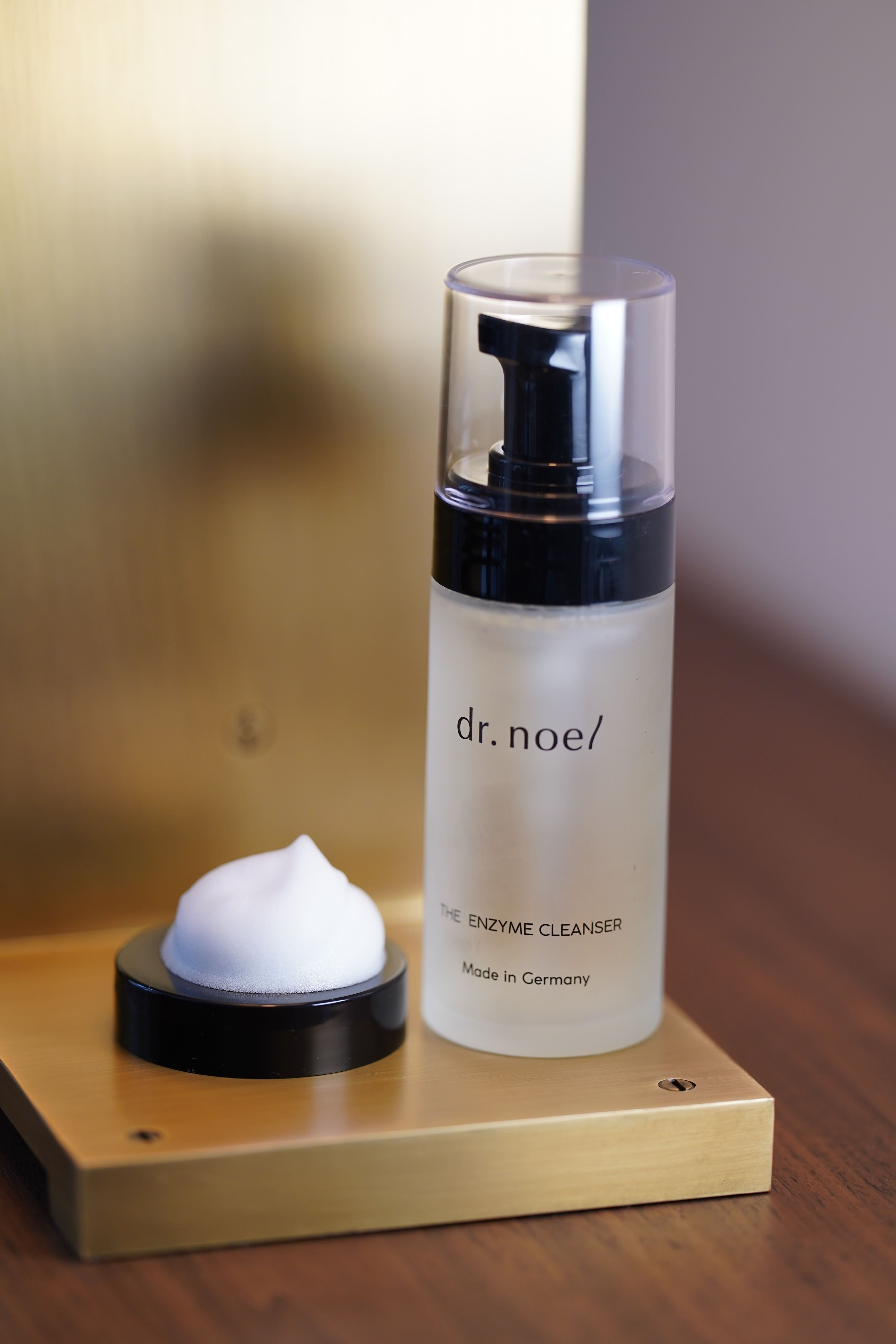 Flasche mit Sprühkopf und Schaum. Aufschrift: dr. noel/ THE ENZYME CLEANSER. Hergestellt in Deutschland. Auf goldenem Podest.