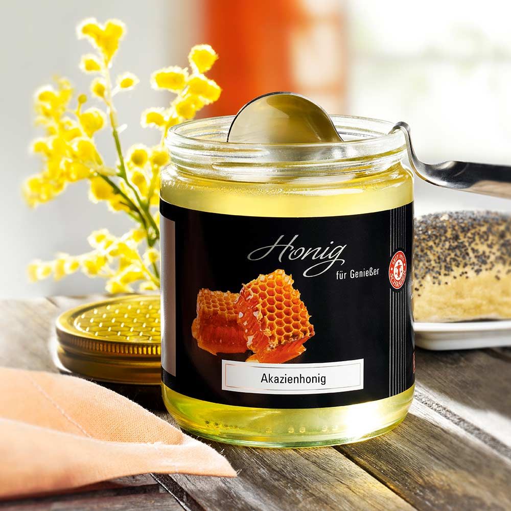 Glas Akazienhonig mit Löffel. Schwarzes Etikett mit Honigwaben-Illustration. Goldener Deckel daneben. Gelbe Blüten im Hintergrund.