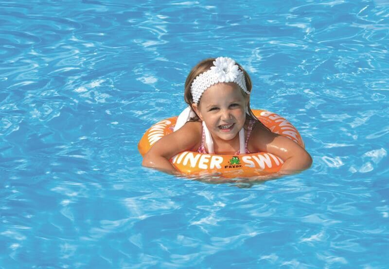 Kind im Wasser mit orangefarbenem Schwimmtrainer. Aufschrift: SWIMTRAINER. Kind lächelt.