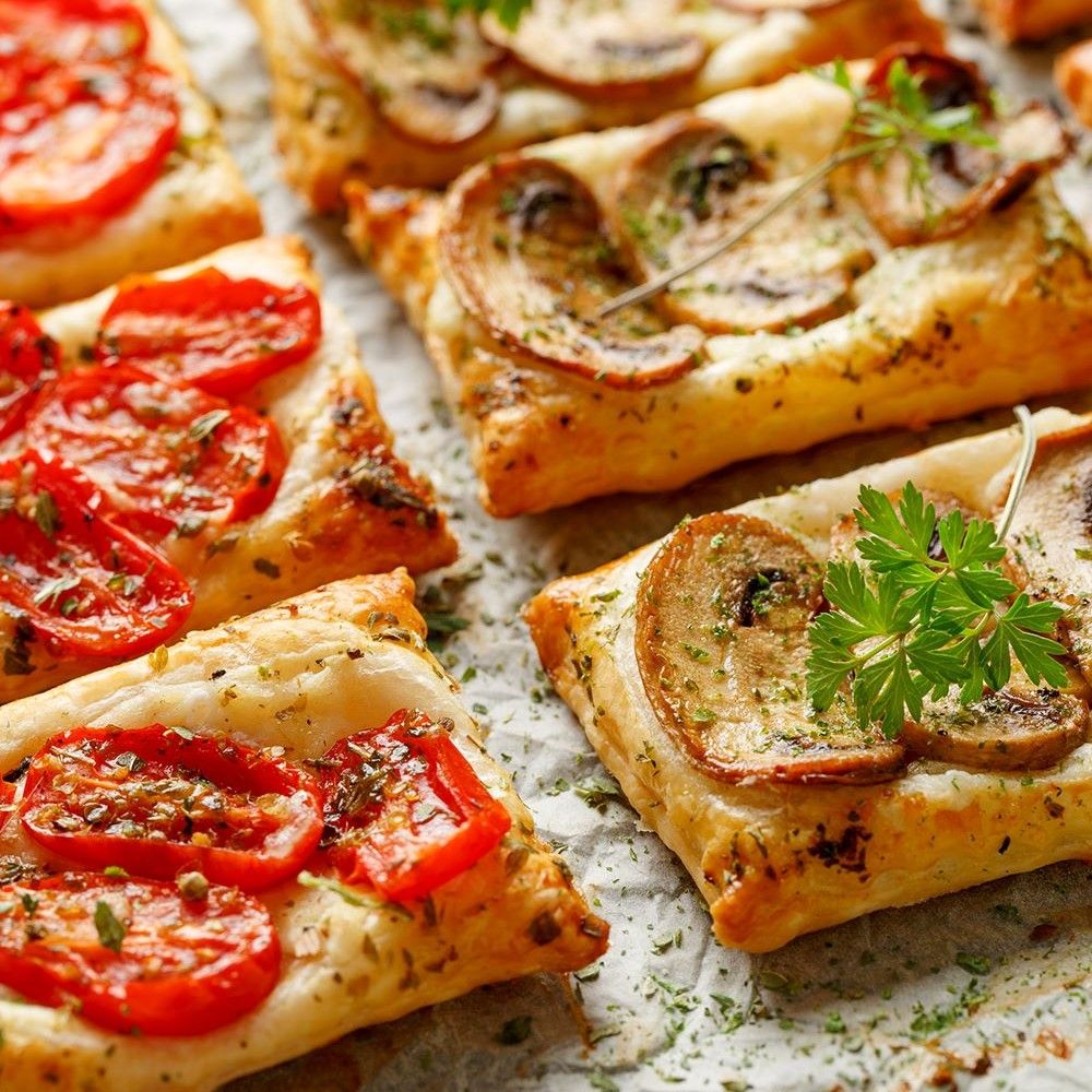 Gebäckstücke mit Tomaten und Pilzen belegt. Quadratische Form, goldbraun gebacken. Dekoriert mit Kräutern.